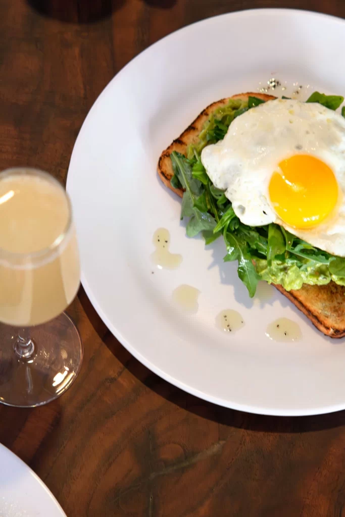 A plate of avocado toast topped with a poached egg alongside a chilled glass of mimosa for brunch.